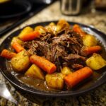 A close-up of a plate filled with tender slow cooker pot roast, chunks of potatoes, and carrots in a rich gravy.