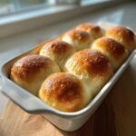 A white baking dish filled with eight golden-brown, shiny sourdough discard rolls, perfectly baked and soft.