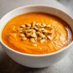 Close-up of a vibrant orange bowl of spicy pumpkin soup garnished with toasted pumpkin seeds.