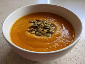 Close-up of a vibrant orange bowl of spicy pumpkin soup garnished with toasted pumpkin seeds (pepitas).