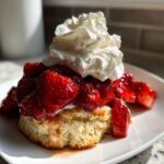 Close-up of a flaky biscuit shortcake topped with macerated strawberries and a dollop of whipped cream.