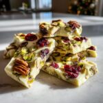 Close-up of white chocolate pretzel bark pieces topped with pistachios, pecans, and dried cranberries.