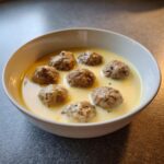 A white bowl filled with youvarlakia Greek meatball soup, featuring seven meatballs in a creamy, pale yellow avgolemono sauce.