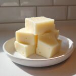 A small stack of creamy, pale yellow white chocolate fudge cubes resting on a white saucer.