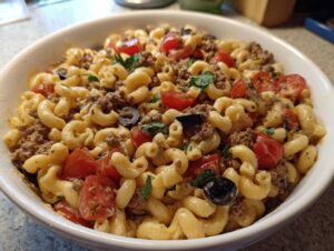 A close-up view of a white bowl filled with creamy taco pasta salad featuring elbow macaroni, ground beef, tomatoes, and black olives.