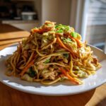 A generous serving of homemade chicken chow mein recipe piled high on a white plate, featuring noodles, chicken, carrots, and cabbage.