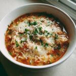 A close-up of a bowl of rich, tomato-based chicken parmesan soup topped with melted mozzarella and fresh parsley.