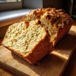 A rustic loaf of golden brown soda bread, with one thick slice cut and displayed on a wooden cutting board.