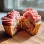 A close-up of amazing strawberry cupcakes cut in half showing pink frosting and fruit pieces inside the cake.