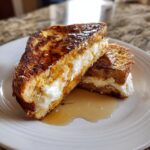 Two halves of golden brown stuffed french toast overflowing with creamy white filling and drizzled with syrup.