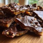 Close-up of broken pieces of homemade toffee recipe, featuring a layer of caramel, nuts, and a dark chocolate topping sprinkled with sea salt.