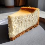 A thick, creamy slice of baked cheesecake recipe with a golden top and graham cracker crust.