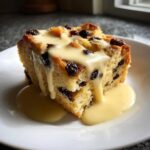 A thick, golden slice of raisin bread pudding generously topped with flowing vanilla custard sauce.