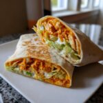 Close-up of a buffalo chicken wrap cut in half, showing shredded chicken coated in orange sauce, lettuce, and ranch dressing.