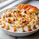 A creamy white apple dip topped with caramel drizzle and crunchy bits, served with fresh apple slices.