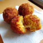 Three golden-brown, deep-fried rice balls, one broken open revealing melted cheese inside. Perfect cheesy rice balls.