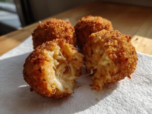 Four golden-brown, crispy rice balls resting on a white napkin, one broken open showing melted cheese stretching inside the cheesy rice balls.