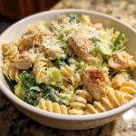 Close-up of a white bowl filled with chicken caesar pasta salad, featuring rotini pasta, grilled chicken chunks, romaine lettuce, and grated Parmesan.