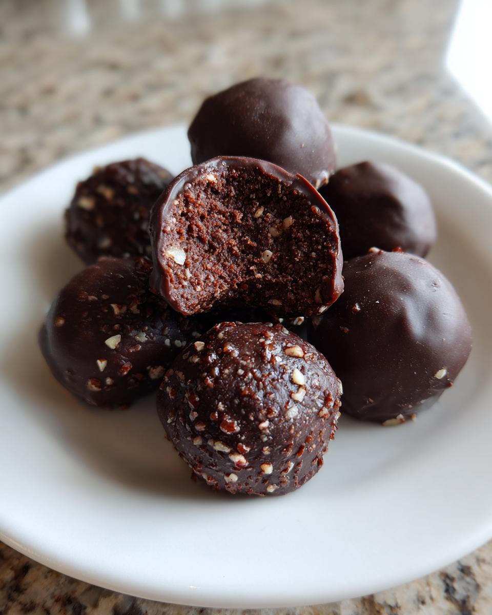 A stack of rich, dark chocolate pecan pie balls on a white plate, one is bitten showing the dense, nutty interior.