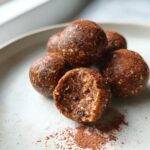 A stack of rich, brown protein balls dusted with cocoa powder, one is bitten to show the texture inside.