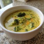 Close-up of a white bowl filled with creamy, yellow broccoli cheddar soup topped with fresh broccoli florets.