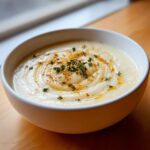 A close-up of a creamy white garlic soup in a white bowl, garnished with thyme sprigs and an oil swirl.