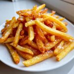 A close-up of a pile of golden, crispy air fryer french fries sprinkled generously with coarse sea salt on a white plate.