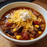 A close-up of a bowl of rich crockpot cowboy soup topped with shredded cheddar cheese and a dollop of sour cream.