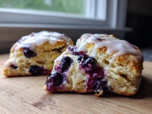 Close-up of two flaky blueberry scones, one cut in half showing juicy berries, topped with a sweet white glaze.