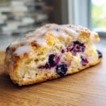 A close-up shot of a flaky blueberry scone topped with white glaze, showing juicy blueberries inside.