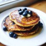 A stack of three golden brown almond flour pancakes topped with butter, blueberries, and syrup.