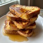A tall stack of golden brown french toast slices generously drizzled with maple syrup on a white plate.