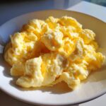A close-up of perfectly cooked, fluffy scrambled eggs served on a white plate, highlighted by bright sunlight.