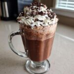 A tall glass mug filled with rich, blended frozen hot chocolate, topped with whipped cream and chocolate shavings.