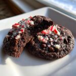 Close-up of fudgy chocolate peppermint cookies, one broken to show the soft interior, topped with crushed candy canes.
