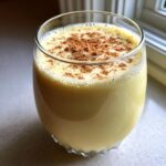 Close-up of a glass filled with creamy, yellow homemade eggnog, dusted generously with ground cinnamon.