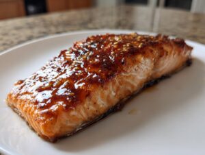 Close-up of a perfectly cooked salmon fillet with a shiny, dark glaze and sesame seeds on a white plate.