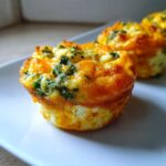 Close-up of a freshly baked, golden brown egg muffin topped with melted cheese and green spinach pieces.