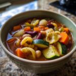 Close-up of a steaming bowl of minestrone soup filled with zucchini, carrots, beans, and shell pasta.