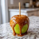 A vibrant green apple fully coated in thick, glossy caramel sauce, creating a perfect caramel apple.