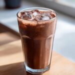 A tall glass filled with iced chocolate milk, showing condensation and ice cubes on top.