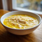 A close-up of a white bowl filled with bright yellow lemon chicken soup, featuring shredded chicken and orzo pasta.