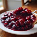 A spoonful of thick, glossy cherry pie filling being lifted from a plate of pie, showcasing the rich color of the cherry pie filling.