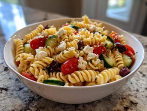 A close-up of a white bowl filled with vibrant mediterranean pasta salad featuring rotini pasta, tomatoes, cucumbers, olives, and feta cheese.