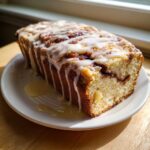 A loaf of moist apple fritter bread, drizzled heavily with white vanilla icing, resting on a white plate.