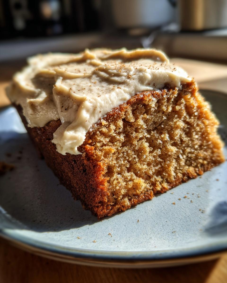 Close-up of a moist slice of applesauce cake topped with creamy frosting and a sprinkle of spice.