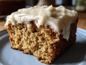 Close-up of a moist slice of applesauce cake topped with thick, creamy frosting dusted with spice.