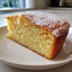 Close-up of a moist slice of yellow olive oil cake dusted with powdered sugar on a white plate.