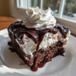 A moist slice of chocolate poke cake topped with white whipped cream and drizzled with rich chocolate syrup.