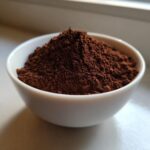 A small white bowl filled with a mound of dark brown, finely ground powder, representing homemade hot cocoa mix.